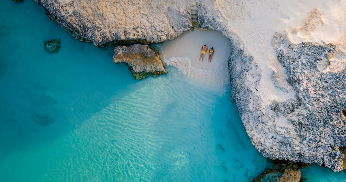 Aerial view of a couple relaxing on a secluded white-sand beach beside turquoise water, representing where private aviation can take you